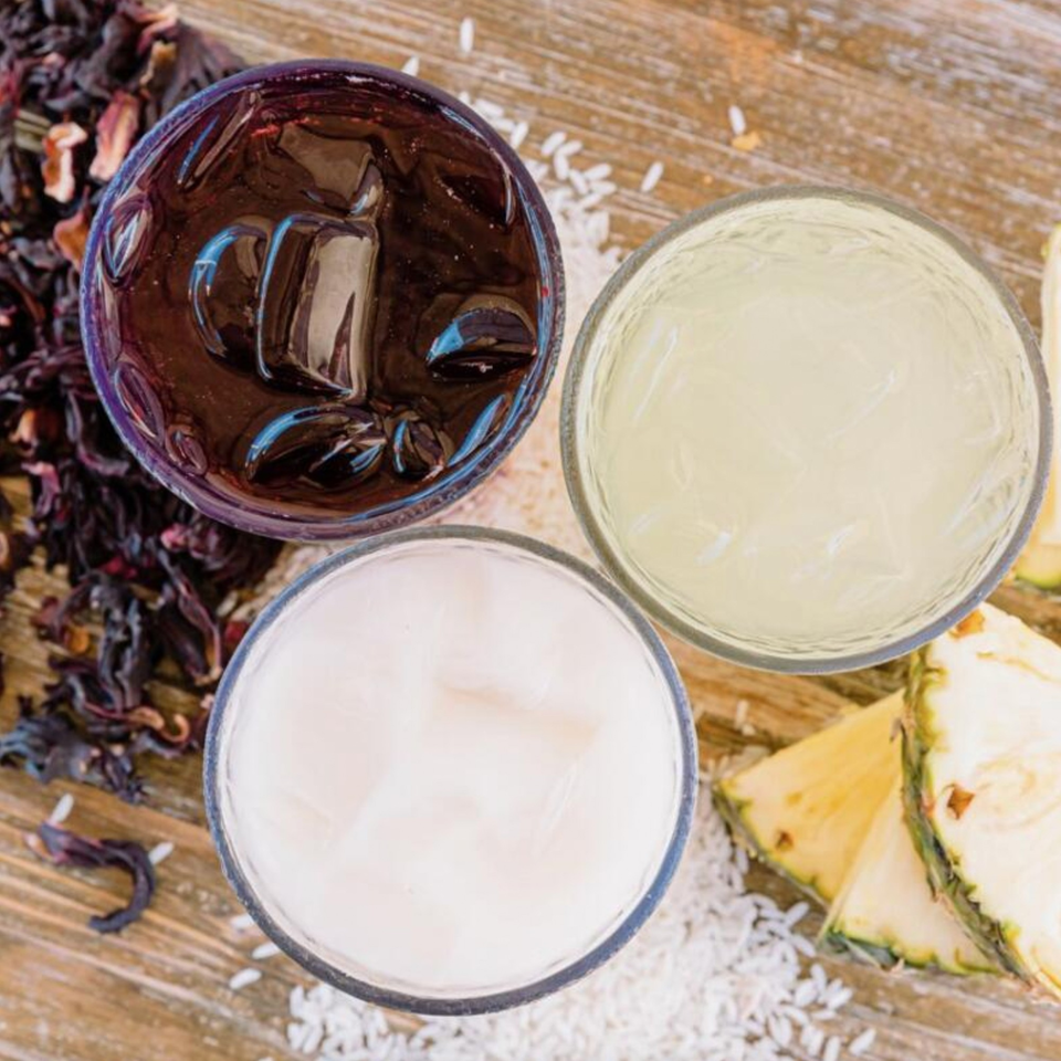 Three glasses of aguas frescas, including jamaica, horchata, and piña, on a wooden surface.