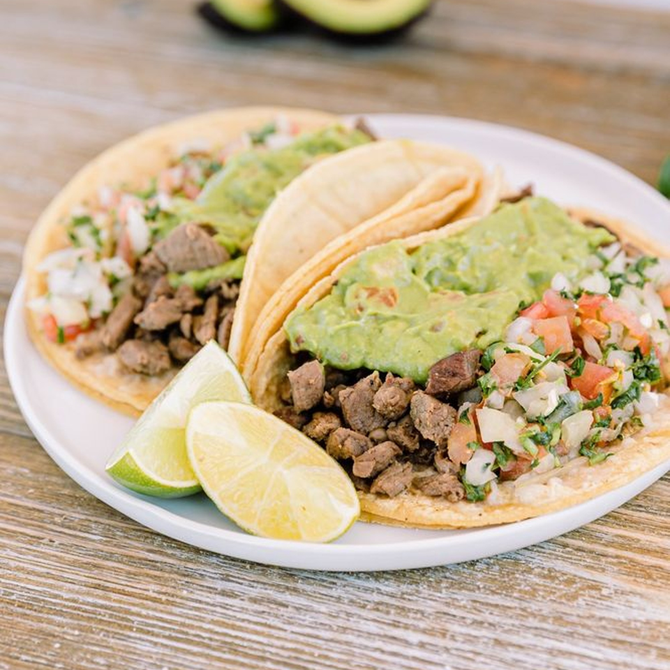 Three Adalbertos Mexican Food tacos with carne asada, guacamole, pico de gallo, and lime wedges.